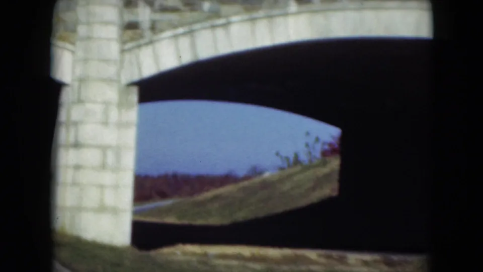 The car passes under the stone arch bridge, with the dark underside framing a grassy slope, pale masonry, and distant tree line.