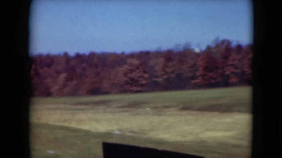 1975 8mm rural Pennsylvania view of open grassland and a wooded horizon with reddish-brown autumn foliage.