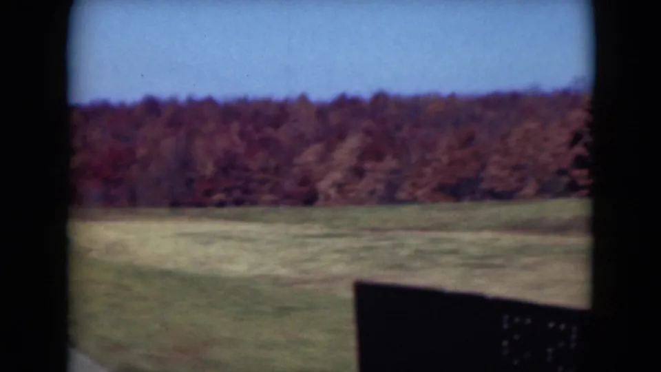 1975 8mm frame from Middletown, Pennsylvania, showing a broad grassy field and dense autumn woods from a moving vehicle.