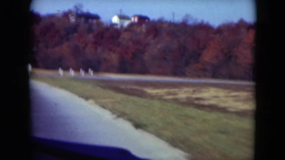 1975 8mm roadside view near Middletown with a paved shoulder, white markers, grassy verge, and autumn woods.
