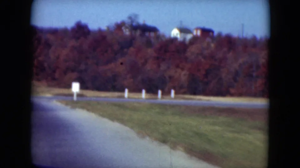 Final 1975 Middletown-area frame showing roadside posts, open grass, red-brown woods, and distant houses.