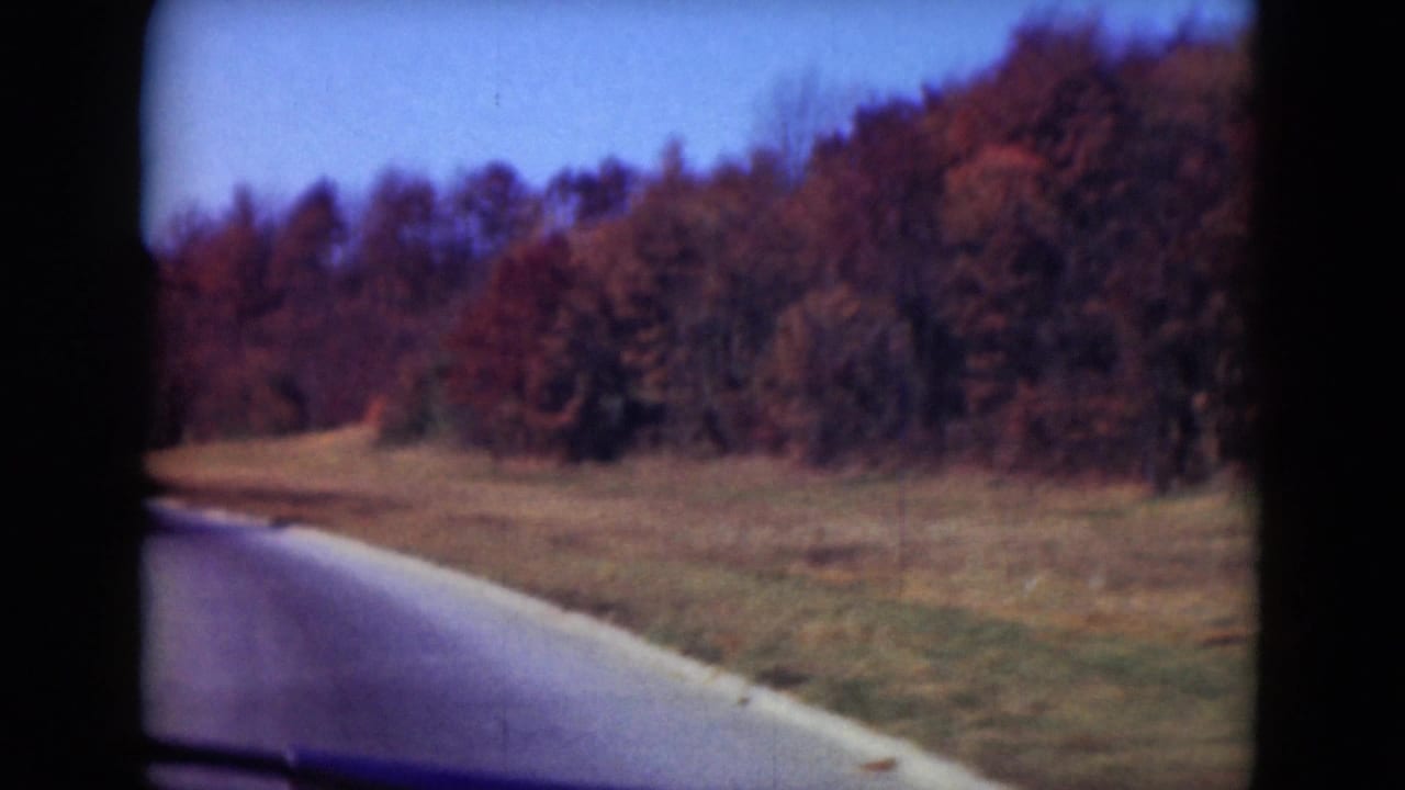 Moving-car view in Middletown, Pennsylvania, with a road bending beside open grass and a tree-covered hillside under blue sky.