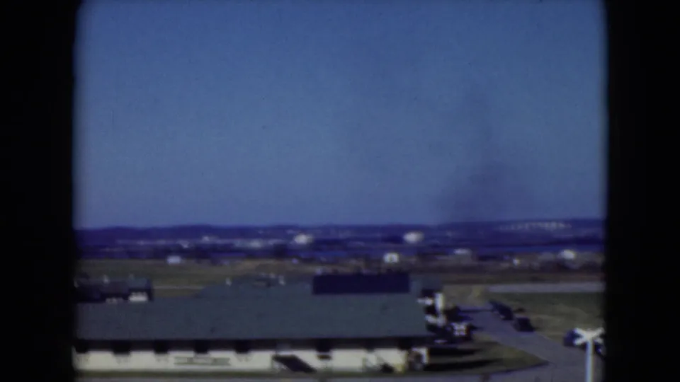 1975 Middletown frame showing a low building, paved road, distant structures, and a faint smoke plume on the horizon.
