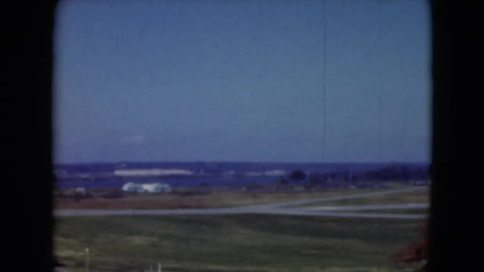 1975 8mm view near Middletown, Pennsylvania with open fields, a curving road, distant structures, and a low horizon beneath blue sky.