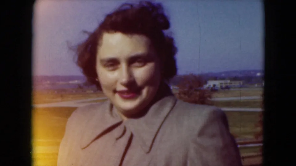 Young woman in a gray coat smiles toward the camera outdoors, with open Pennsylvania fields behind her in 1975 8mm footage.