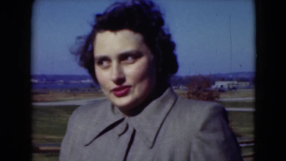 Woman in a gray coat faces the camera near open fields and a distant vehicle in 1975 Middletown, Pennsylvania home-movie footage.