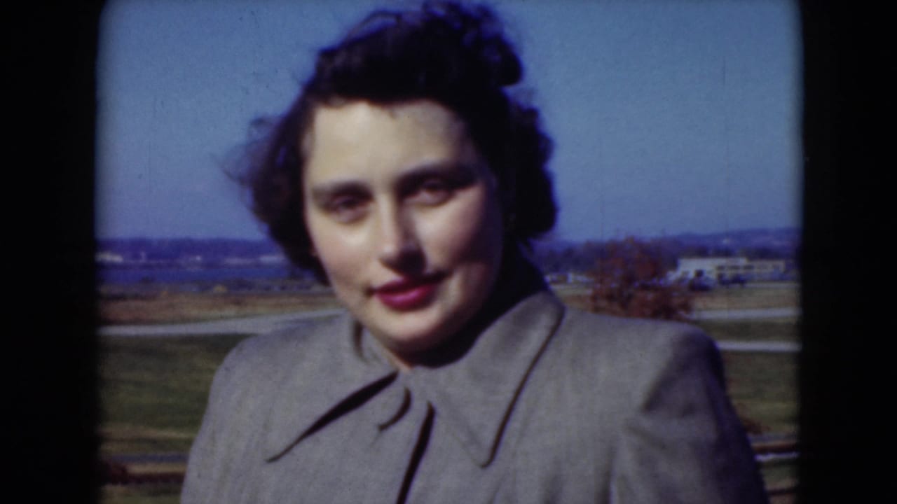Dark-haired woman in a gray coat standing outdoors with fields behind her.