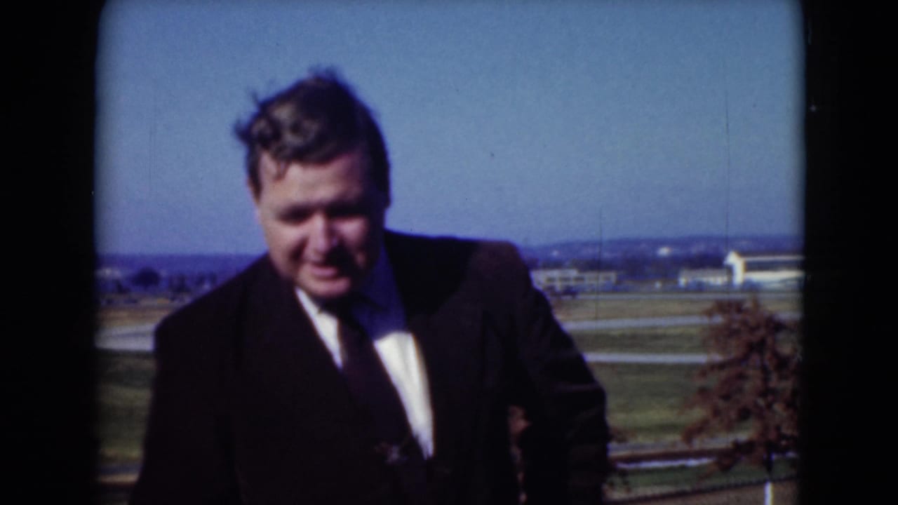 Man in a dark suit and tie stands outdoors in the 1975 Middletown home-movie scene, with fields, trees, and low buildings in the distance.