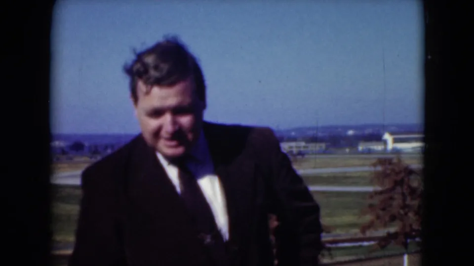 Man in a dark suit and tie stands outdoors in the 1975 Middletown home-movie scene, with fields, trees, and low buildings in the distance.
