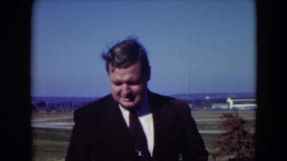 The suited man looks down or smiles slightly during the Middletown outdoor home-movie moment, framed by fields and a clear sky.