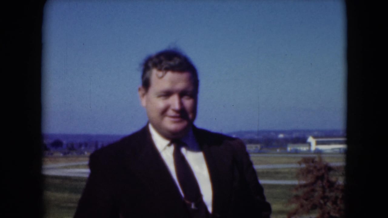 Man in dark suit and tie standing outdoors near a road with distant buildings behind him.