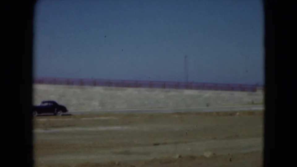 A dark older car passes along the Middletown roadside, with a concrete wall, red railing, and smokestacks in the background.