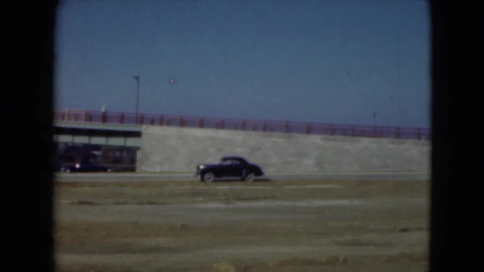 1975 8mm roadside frame showing a black car, another vehicle, and a small structure near the Middletown aqueduct or embankment.