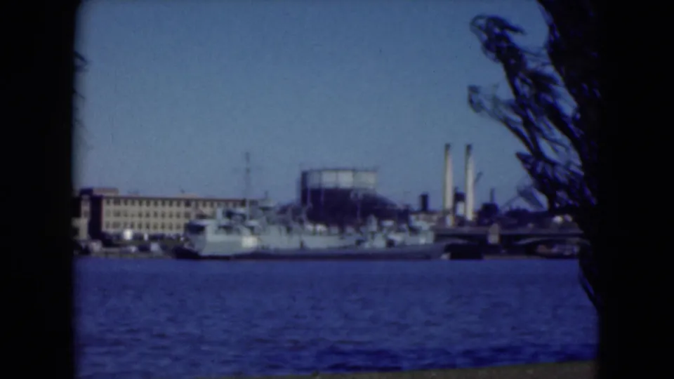 1975 8mm waterfront view in the Middletown, Pennsylvania clip, with a docked naval ship, industrial buildings, smokestacks, and water.