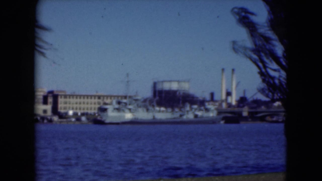 Naval ship on a river near industrial buildings, framed by dark trees.