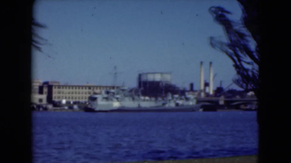1975 Middletown, Pennsylvania 8mm frame showing a naval ship moored along a riverfront with buildings and smokestacks behind it.