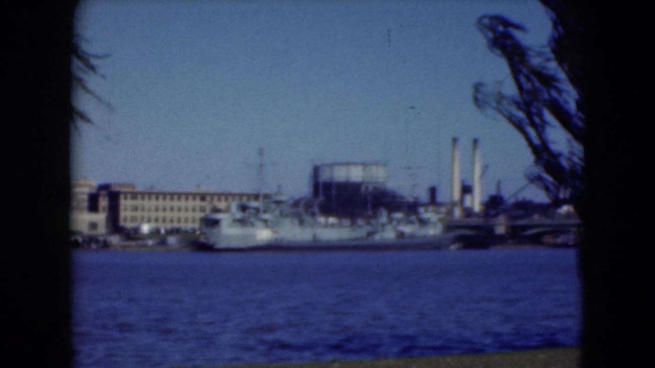 Riverfront view of a naval ship, buildings, smokestacks, and tree silhouettes.