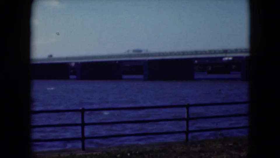 1975 Middletown, Pennsylvania 8mm frame showing a bridge across the river, with a railing and tree trunks in the foreground.