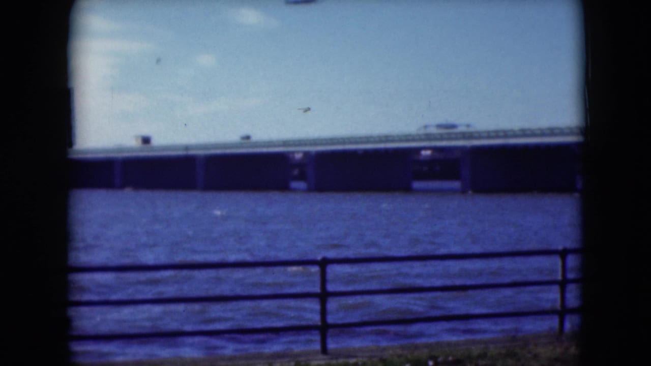 Long bridge over blue water seen past a metal railing, with a dark film border at the edge.