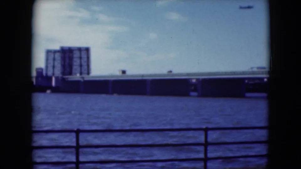 1975 Middletown, Pennsylvania, home-movie frame with a bridge, blue water, railing, and large industrial building across the river.