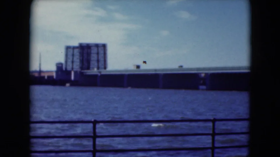 1975 8mm frame of a Middletown, Pennsylvania, waterfront view with bridge infrastructure and a large industrial structure beyond the water.