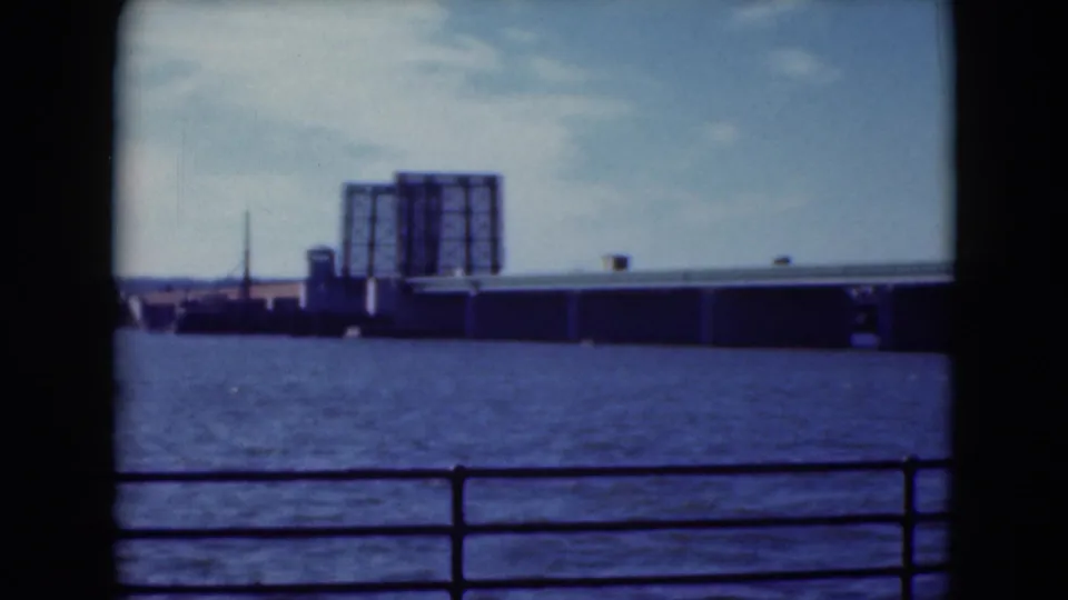 1975 Middletown, Pennsylvania, 8mm frame showing a long bridge over blue water, a foreground railing, and industrial buildings on the far shore.