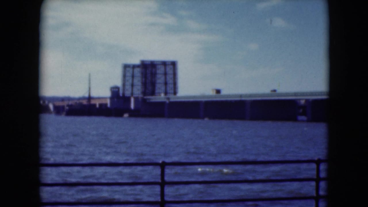 1975 8mm travel frame showing a waterfront railing, open water, and a distant bridge or industrial shoreline structure.