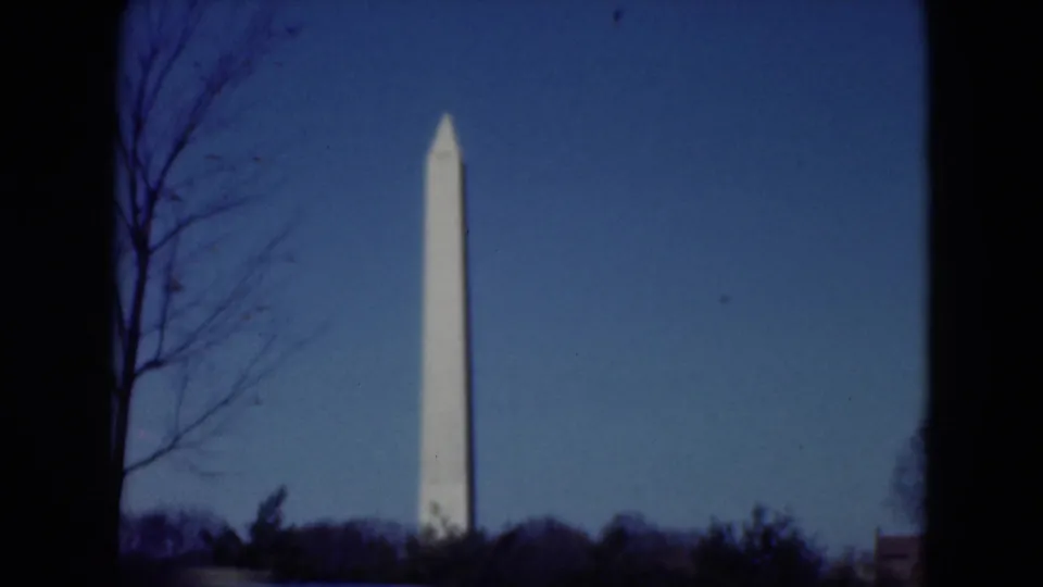 A distant white obelisk stands against a blue sky, partly framed by bare trees and dark film-edge shadows in 1970s 8mm footage.