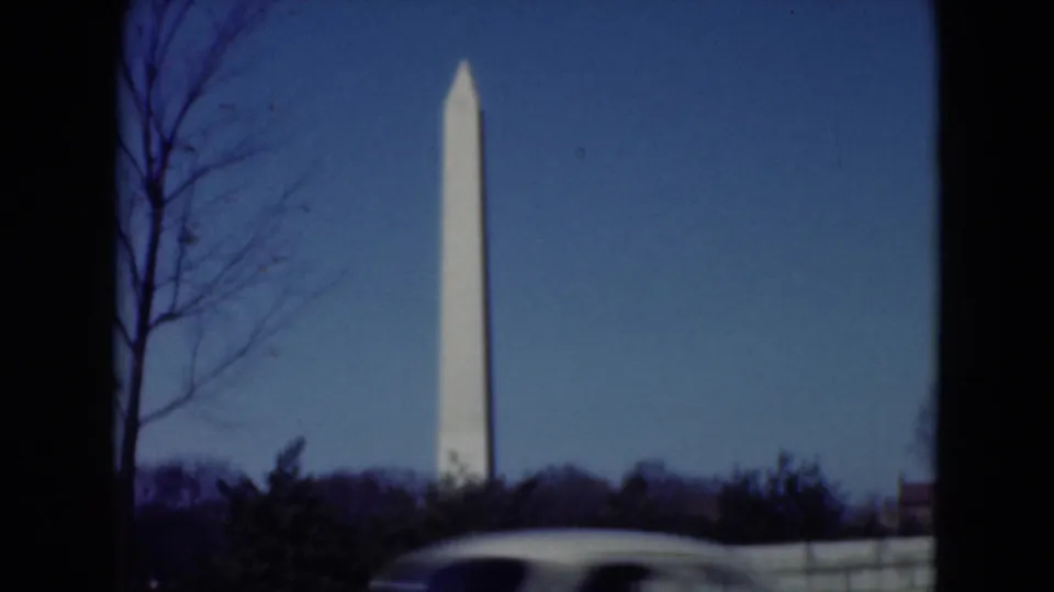 The obelisk remains centered beyond winter trees as a light-colored car passes through the foreground of the home-movie frame.