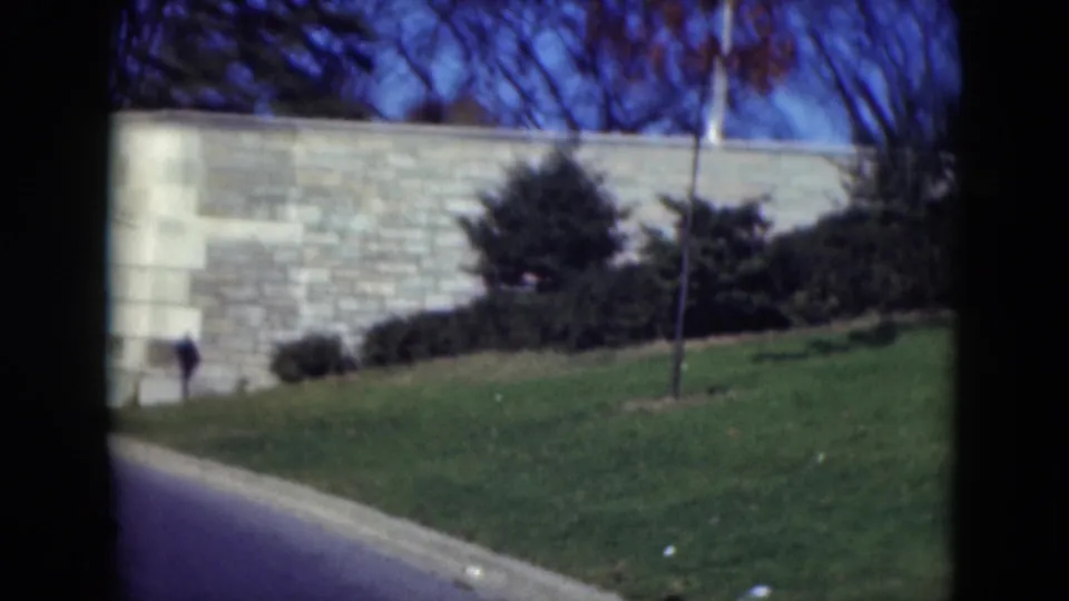 The view shifts to a pale stone wall, grass, shrubs, and bare trees, with only a partial glimpse of the obelisk near the upper edge.