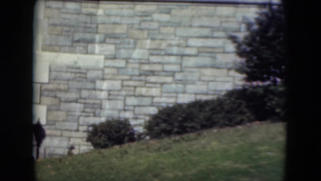 Close view of a stone wall with shrubs, grass, a tree edge, and a small distant figure near the center.