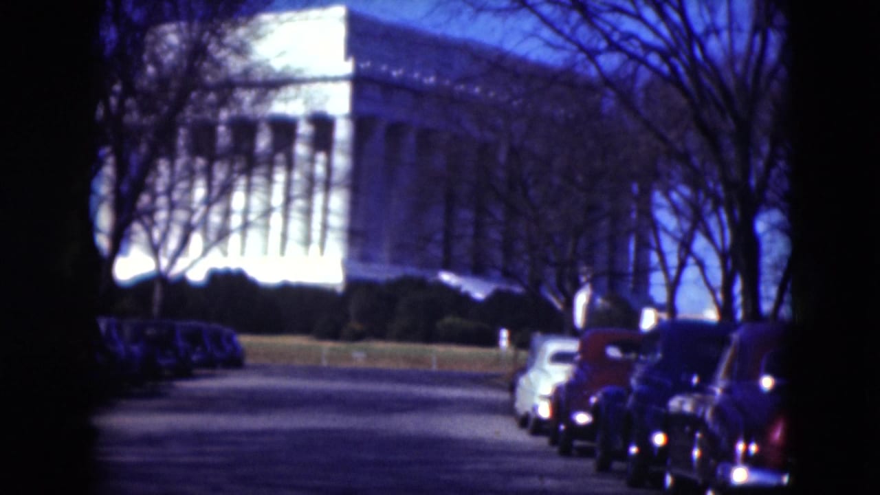 The camera is closer to the white columned building, with parked cars on the roadside and leafless branches crossing the view.
