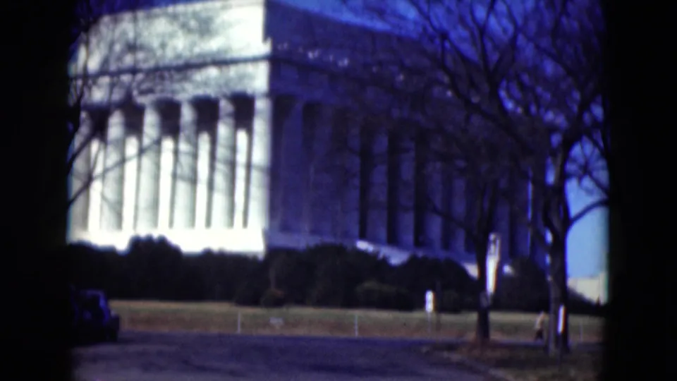 A closer 8mm view of the large white building shows tall columns, dark shrubs, bare trees, lawn, and a roadside foreground.
