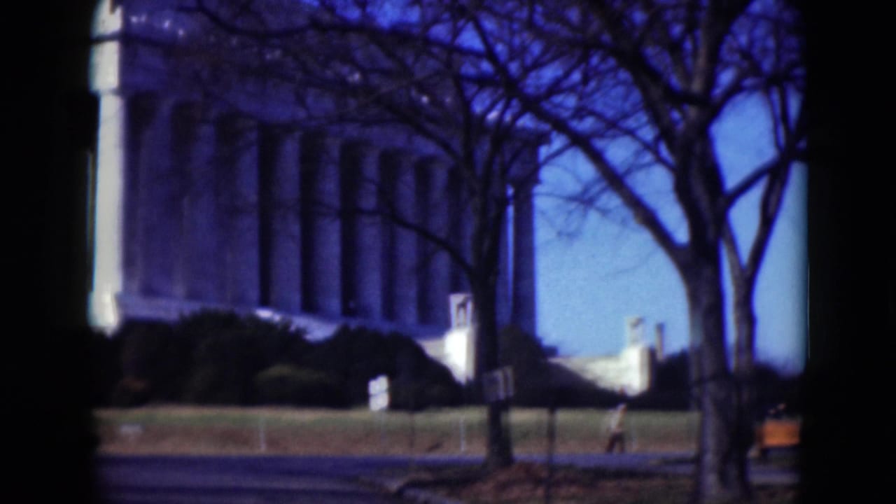 1970s 8mm view of the Lincoln Memorial area through dark foreground framing, with bare trees and bright blue sky.