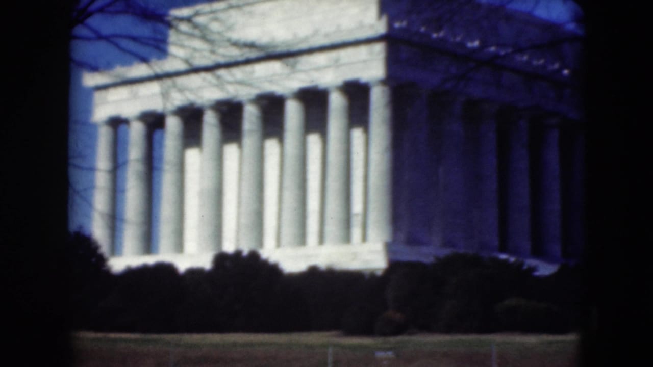 Closer 8mm view of the Lincoln Memorial facade, framed by dark foreground edges, bare branches, and low shrubs.