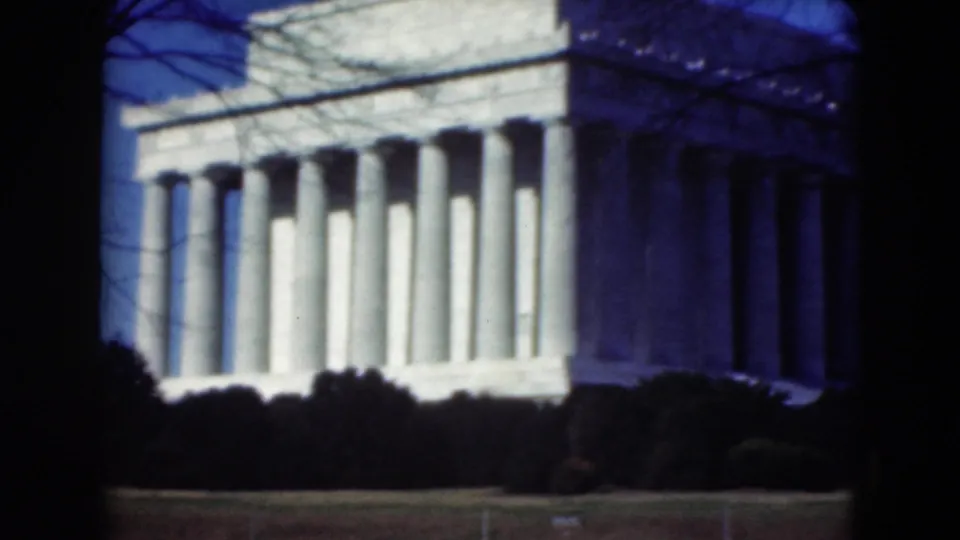 Closer 8mm view of the Lincoln Memorial facade, framed by dark foreground edges, bare branches, and low shrubs.