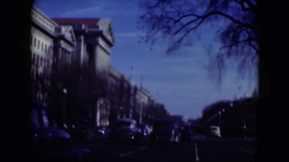 1970s 8mm street view with older cars, bare trees, and large buildings along a broad avenue.