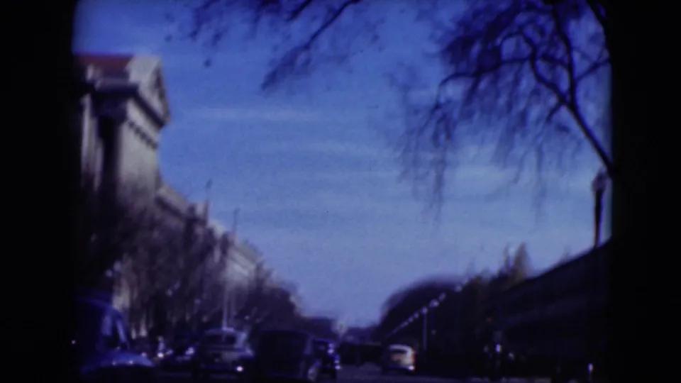 Soft 8mm frame of a broad avenue with cars, bare trees, and buildings, likely filmed from a vehicle.