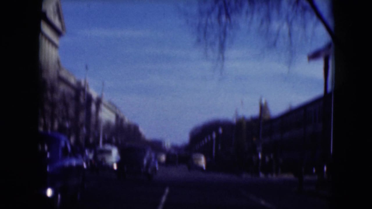 Street seen from a moving vehicle with cars, bare trees, and large buildings in the distance.