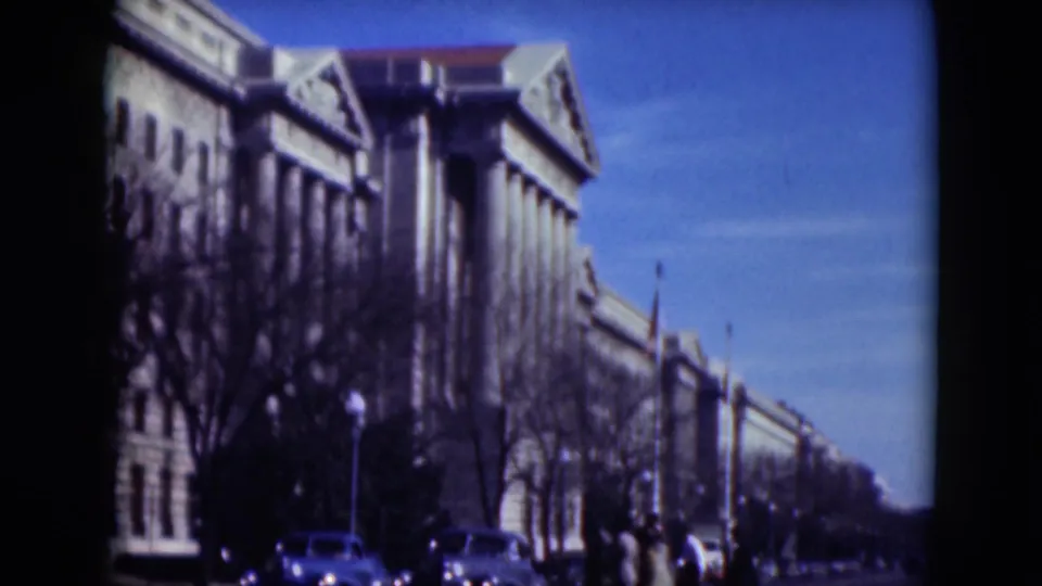 1975 8mm view of a large Middletown building with columns, parked cars, pedestrians, bare trees, and a clear sky.
