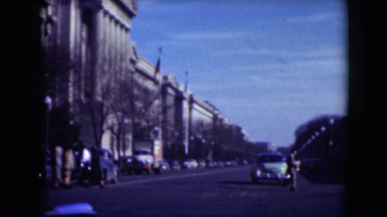 Wide street with cars, pedestrians, and large columned buildings under a blue sky.