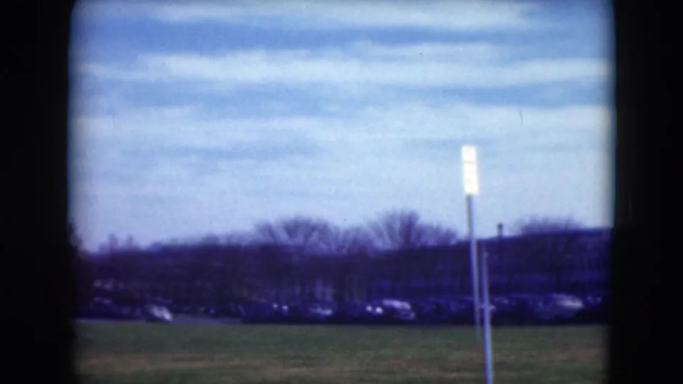 Open grassy field in Middletown with a roadside sign, distant parked cars, low buildings, and leafless trees beneath a pale sky.