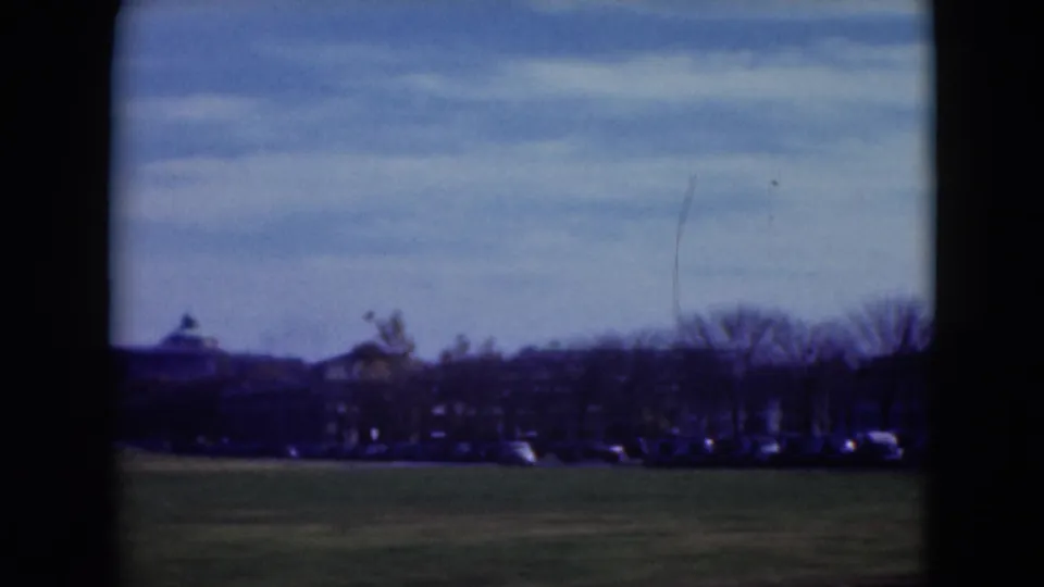 Middletown field view with open grass in the foreground, a row of distant cars, leafless trees, and low structures near the horizon.