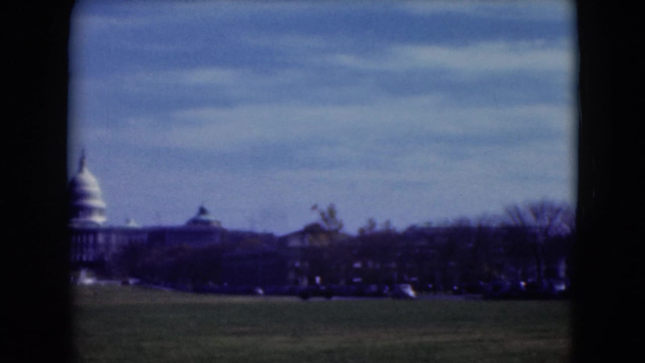 1975 8mm view of the U.S. Capitol seen from a lawn in Washington, D.C., with trees and cars along the horizon.