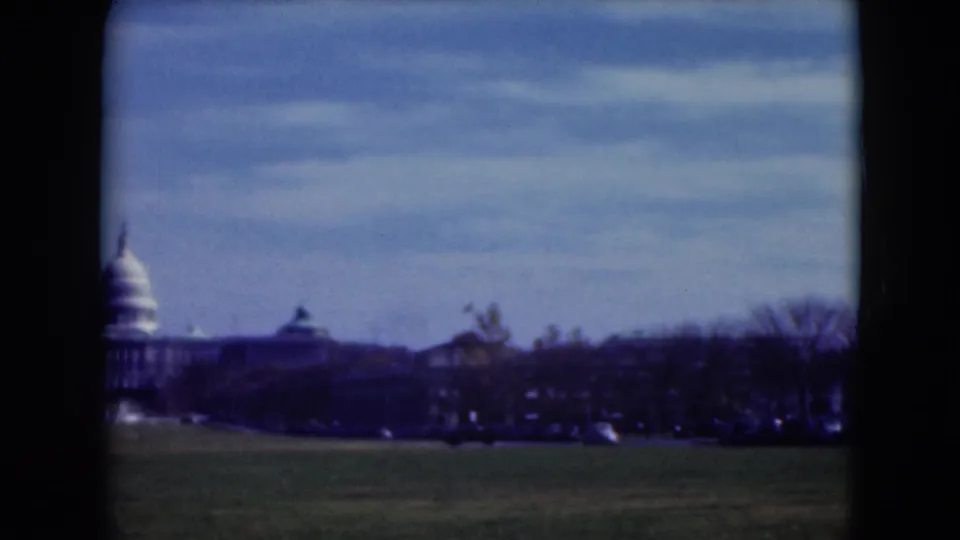 1975 8mm view of the U.S. Capitol seen from a lawn in Washington, D.C., with trees and cars along the horizon.