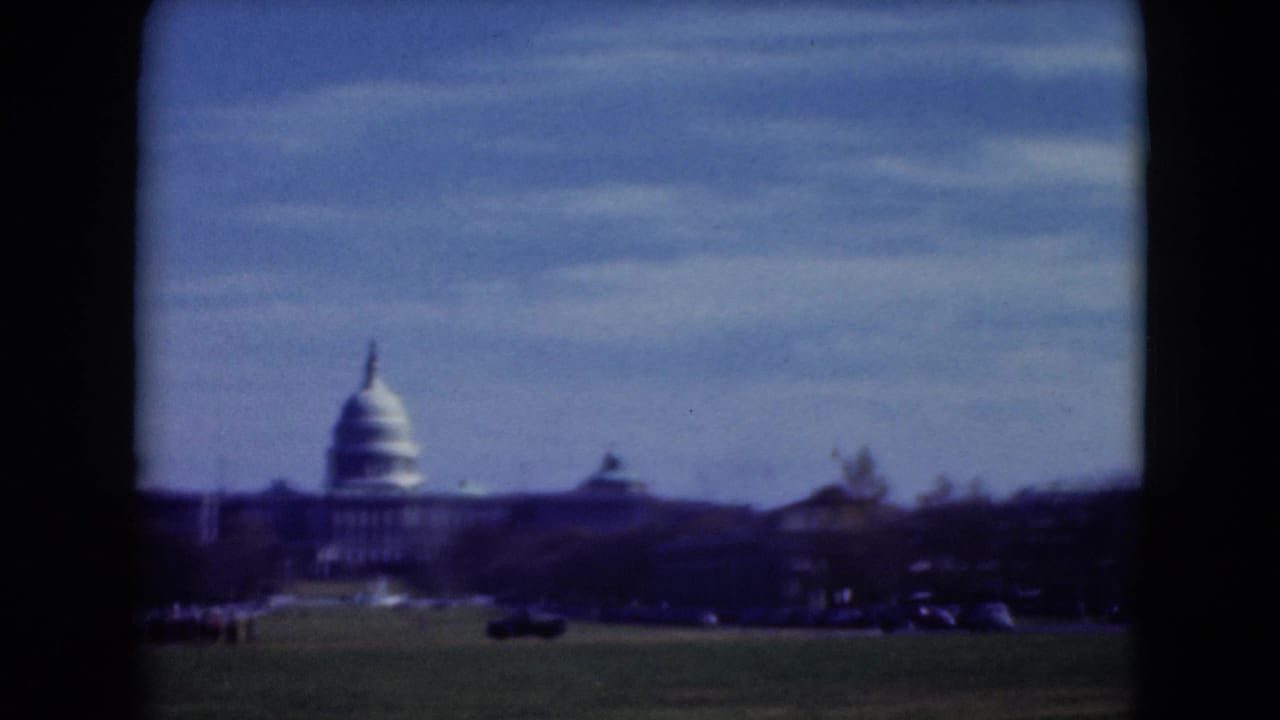 1975 visitor-shot 8mm frame of the U.S. Capitol across open grass, with traffic and trees below a blue sky.