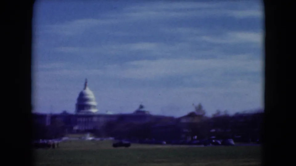 1975 visitor-shot 8mm frame of the U.S. Capitol across open grass, with traffic and trees below a blue sky.