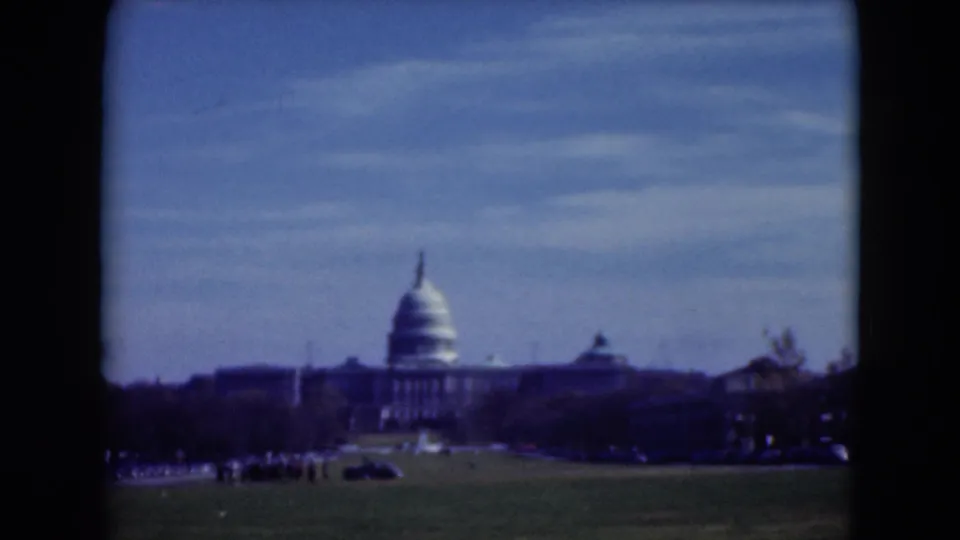 Final 1975 8mm frame of the U.S. Capitol across a green lawn, with distant traffic, trees, and soft blue sky.