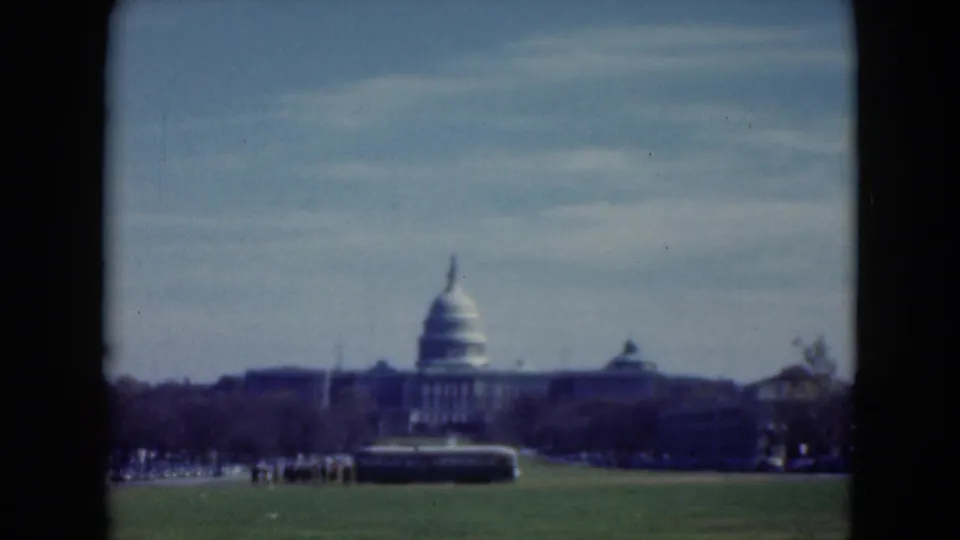 1975 8mm visitor view of the U.S. Capitol dome across a lawn, with a large vehicle and distant people near the trees.