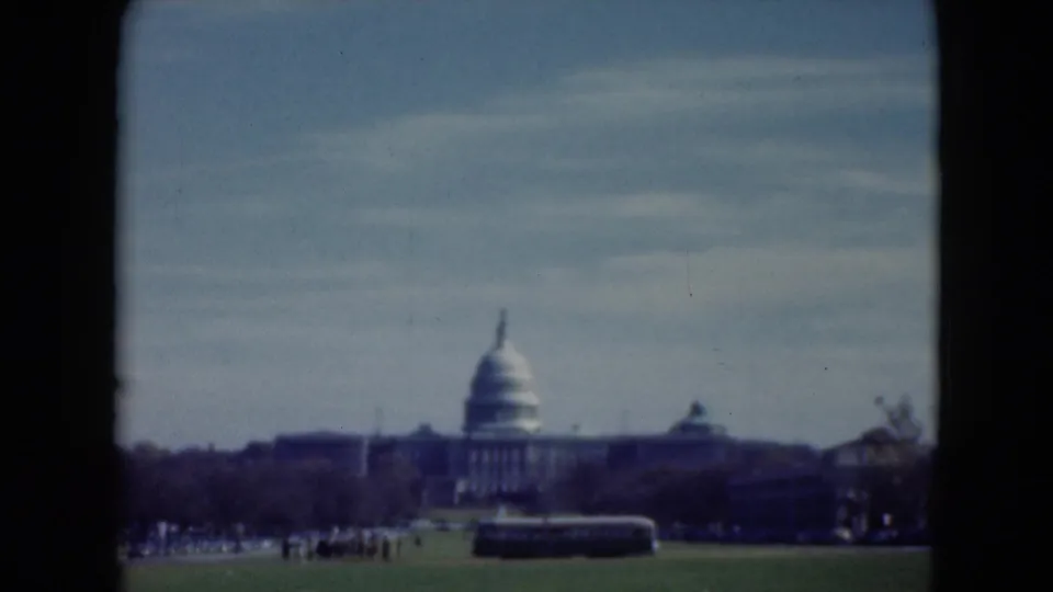 1975 8mm view toward the U.S. Capitol in Washington, D.C., framed by dark film borders with visitors and a bus on the lawn.